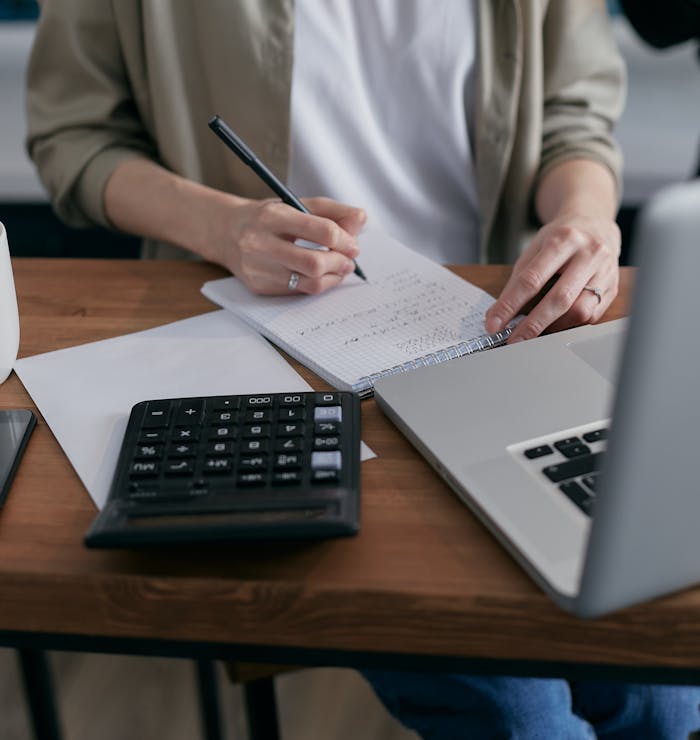 CRA Business Registration Online Changes: What Canadian Businesses Need to Know in 2026 A woman writes financial calculations in a notebook, using a calculator and laptop at a wooden desk.